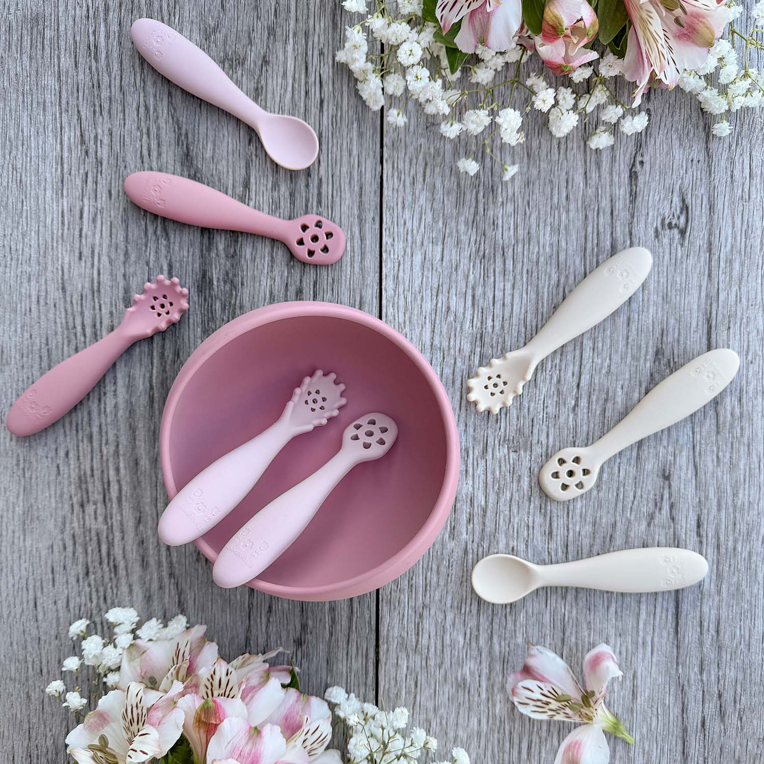 Nybegynnerskjeer i silikon i rosa med dekorative perforerte hoder rundt en matchende skål beige