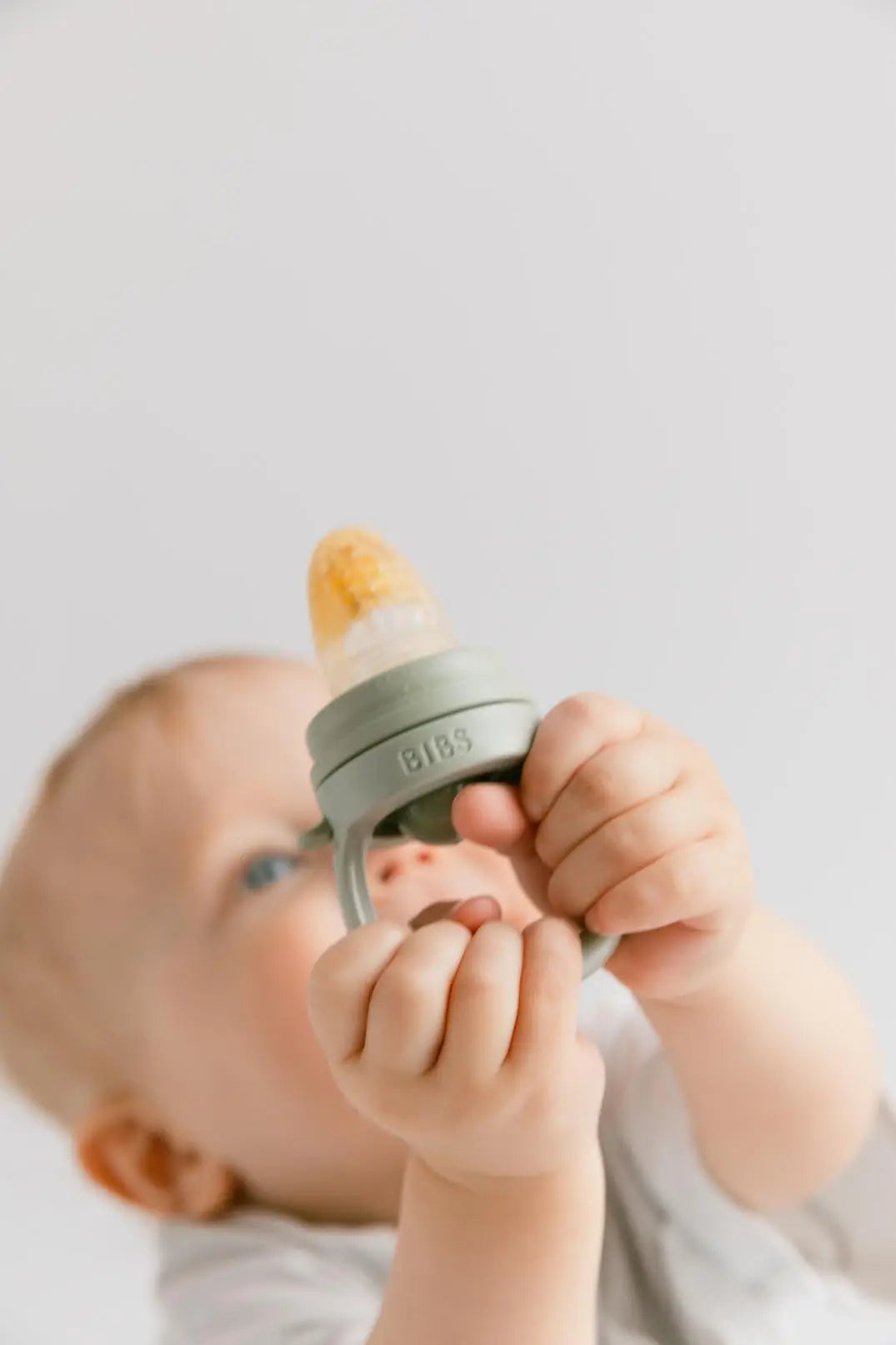 En baby holder et sage grønt silikon smokkesmokk med guloransje tut for baby utforske fast føde