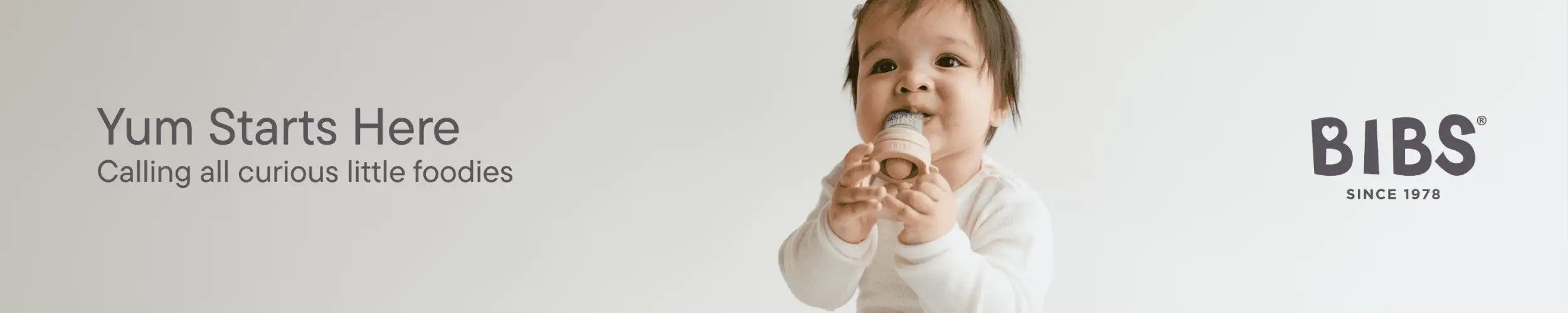 En baby i en hvit skjorte med lange ermer holder en bitleke fra merket bibs, som er laget av myk silikon med en strukturert overflate og har bibs-logoen.