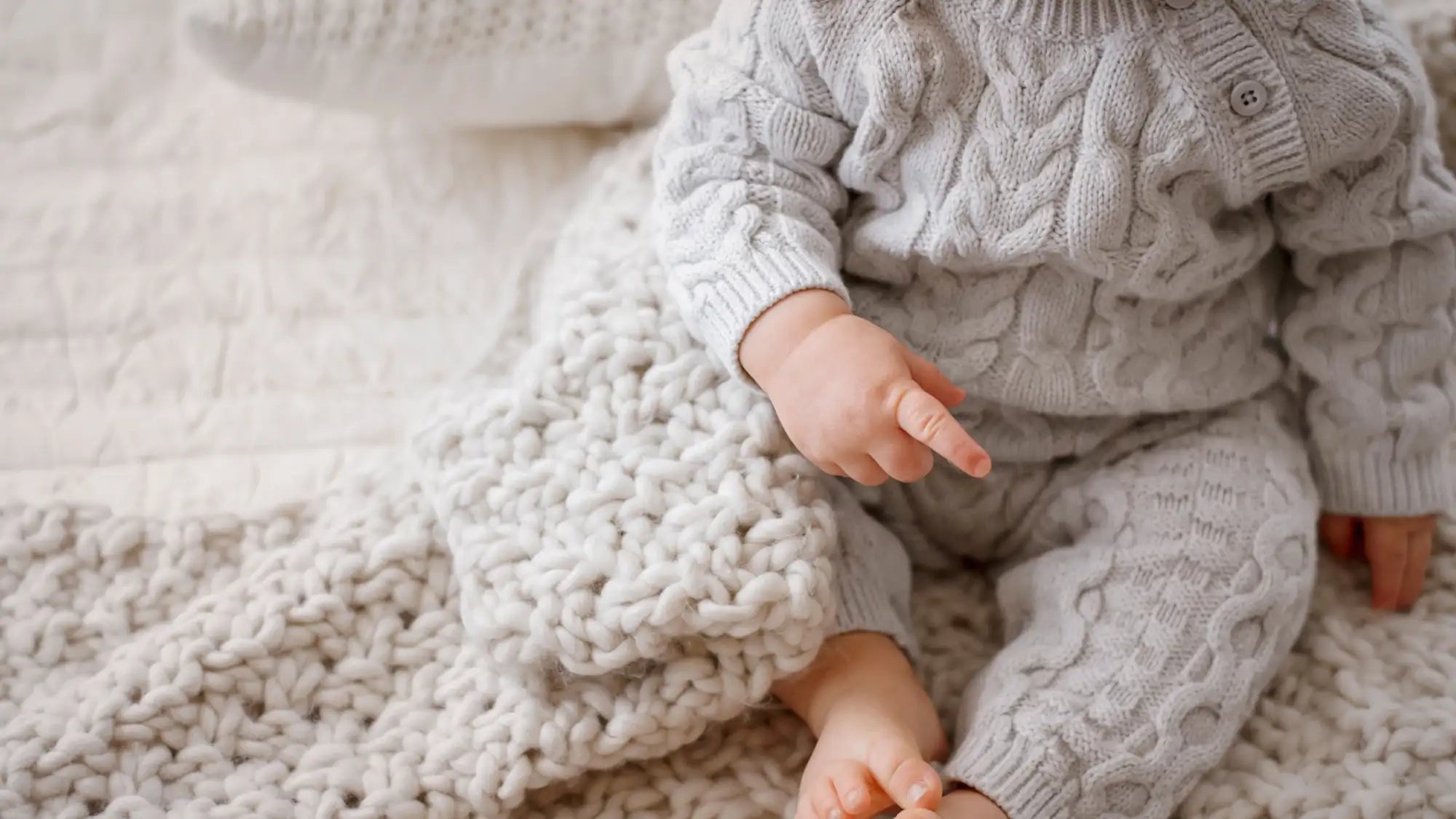En baby som bruker en myk, lys grå kabelstrikket genser med intrikate fletter og matchende bukser.