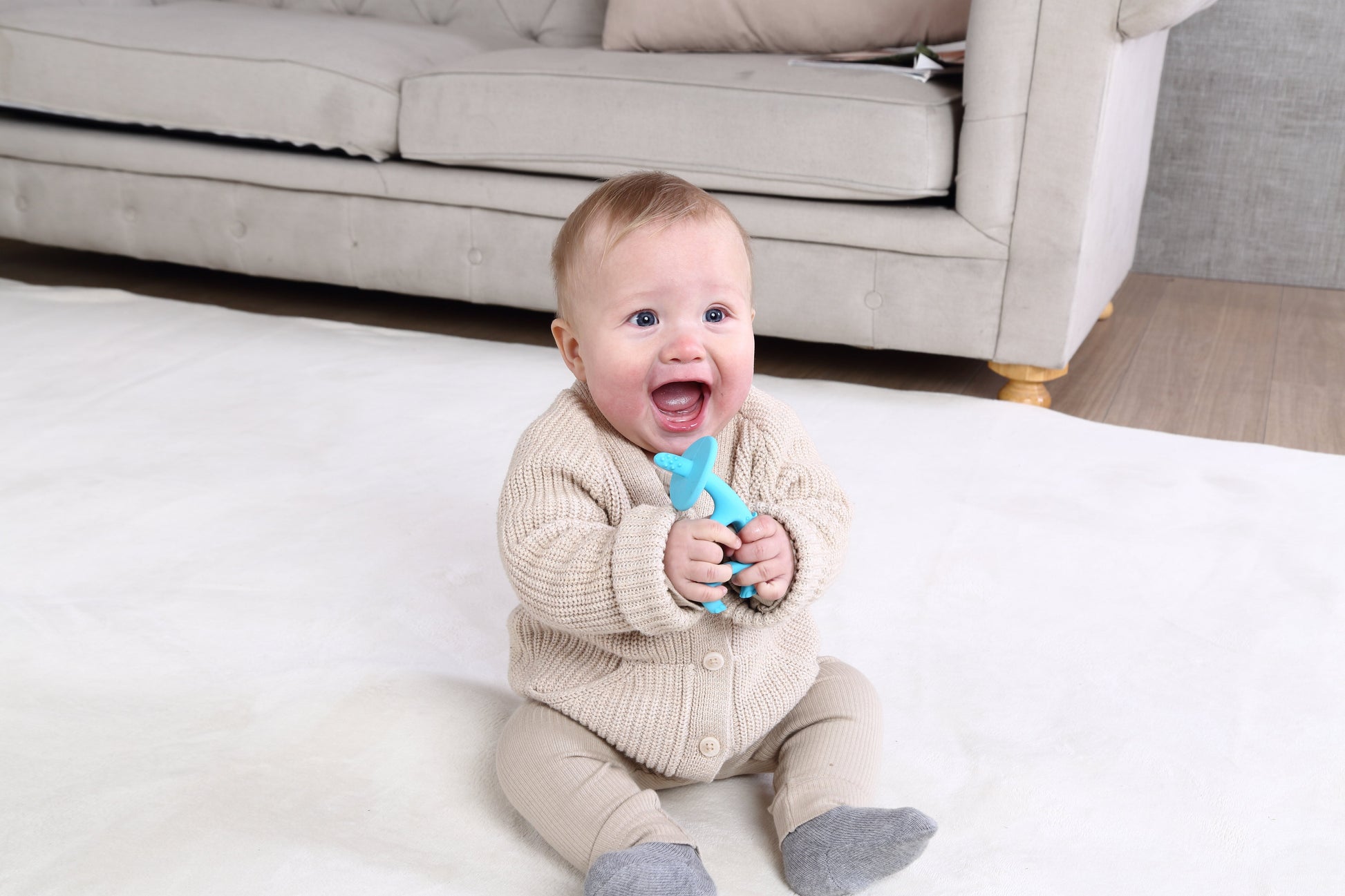En baby i beige strikkejakke holder dansende elefant biteleke blå i silikon som tåler oppvaskmaskin