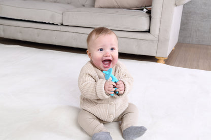 En baby i beige strikkejakke holder dansende elefant biteleke blå i silikon som tåler oppvaskmaskin