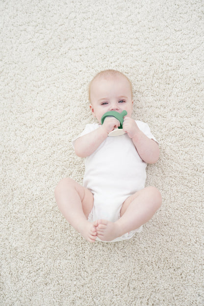 En baby i hvit heldrakt holder en grønnbeige silikon tannutstyr sopp eukalyptus som tåler oppvaskmaskin