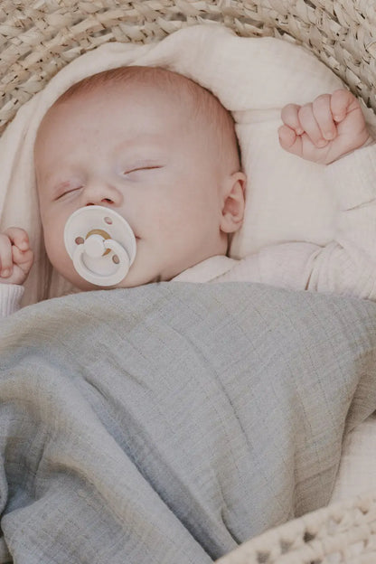 En myk offwhite smokk med subtil tekstur i babyens munn sammen med musselinkluter 70x70 cm babyblå multifunksjonell babyklut